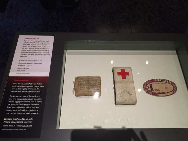 Wording as per the sign on the cabinet - Front-line first aid If you got shot, you prayed a mate would put on your First Field Dressing - just two small wads of gauze, one for the entry would, one for the exit.  Then you hoped like hell a stretcher-bearer would find you in time.  A Red Cross armband was all that stood between those brave men and death.  It showed they were unarmed and shouldn't be targets. First-class care? When Private Joseph Kelly was shot on the first day of the campaign, he was taken back to the troopship Lutzow and this baggage label was tied around his wrist.   The Lutzow - a captured German liner - was so ill-equipped to evacuate casualties that old luggage labels were used to identify the wounded.  The voyage to hospitals in Egypt was a nightmare.  Initially, only two men on board had medical experience: a veterinary surgeon and a medical orderly.